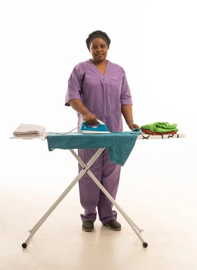 أداء الاعمال المنزلية الروتينية بسعادة ,عاملة منزلية افريقية سمراء البشرة ترتدي يونيفورم بنفسجى اللون تقوم بكي الملابس ,بورتريه خلفية بيضاء بالطول الكامل للجسم