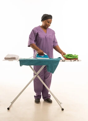 A dark-skinned domestic worker wearing a purple uniform is ironing clothes; a full-length portrait against a white background of an African domestic worker with happy and joyful gestures.