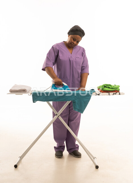 Portrait of a dark-skinned domestic worker wearing a purple uniform while ironing clothes, white background, full-length body.
