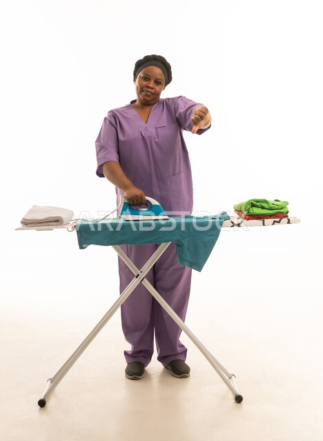 A dark-skinned African domestic worker wearing a purple uniform with her thumb raised in a gesture of disapproval. Portrait with a white background of a domestic worker with a gesture of dissatisfaction.