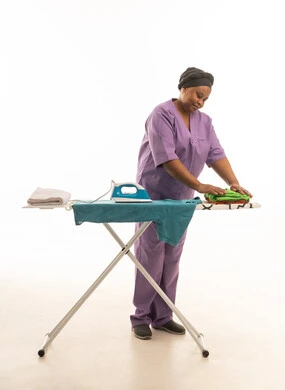 عاملة منزل افريقية ترتدى يونيفورم باللون البنفسجى تقوم بكي و طي الملابس مبتسمة اثناء تأدية عملها  ,بورتريه خلفية بيضاء بالطول الكامل للجسم ,