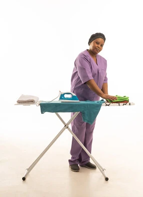Looking at the camera while folding ironed clothes, an African domestic worker wearing a purple uniform with smiling gestures, full-length white background portrait