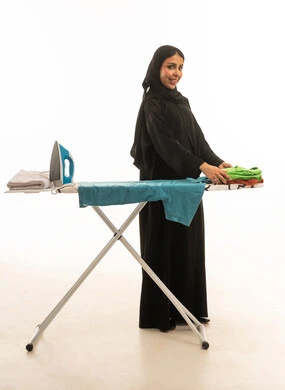 A gesture of happiness and joy from a Saudi Arabian Gulf woman wearing a dark-colored Gulf abaya as she organizes and arranges clothes after ironing. Full-length portrait with a white background and a side view of the camera.