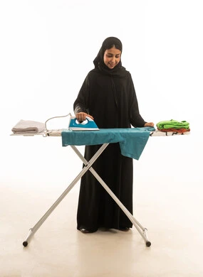 A Saudi Arabian woman from the Gulf region, wearing a black abaya, irons clothes with gestures of happiness and joy. Full-length portrait on a white background.