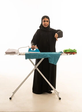 1765971320GaRDkA frowning face with a thumbs-down gesture indicating disapproval. A veiled Saudi Arabian woman wearing a black abaya is ironing and folding clothes. Full-length white background portrait, front view.4Gg4b