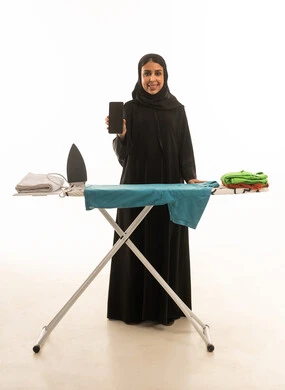 بورتريه خلفية بيضاء للجسم بالطول الكامل لامرأة عربية سعودية خليجية محجبة ترتدي عباءة سوداء , ايماءة سعادة مع عرض شاشة هاتف سوداء فارغة امام طاولة كي الملابس .لقطة أمامية للكاميرا