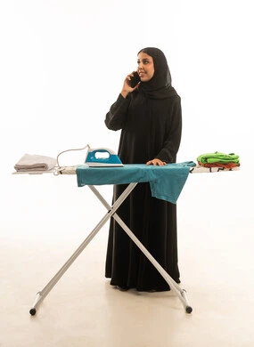 التحدث بالجوال مع كي الملابس وتصفيطها .امرأة عربية سعودية  خليجية محجبة ترتدي عباءة سوداء امام طاولة كي الملابس . بورتريه بالطول الكامل للجسم خليفة بيضاء