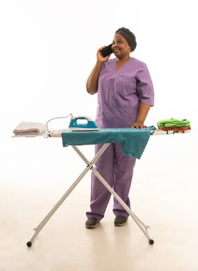 عاملة منزلية افريقية سمراء البشرة ترتدي يونيفورم  باللون البنفسجي تقوم باجراء مكالمة هاتفية مع ابداء السعادة و الابتسامة أثناء أداء المهام المنزلية و كي الملابس وتنظيمها وترتيبها ,بورتريه خليفة بيضاء للجسم بالطول الكامل