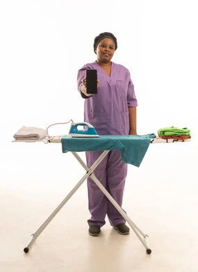 A mobile phone is displayed with a blank black screen facing the camera. A dark-skinned African woman, a domestic worker, is wearing a purple uniform and is ironing and arranging clothes. A full-length portrait of a white mannequin is shown with a front view facing the camera.