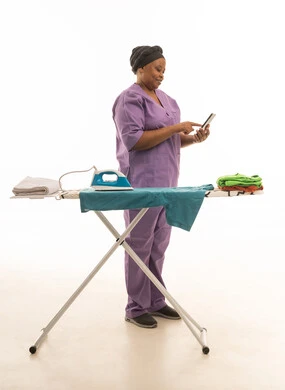 سيدة افريقية سمراء البشرة تقوم بكي الملابس مع ترتيبها وتنظيمها, بورتريه خلفية بيضاء لعاملة منزلية ترتدي يونيفورم   باللون البنفسجي بورتريه للجسم بالطول الكامل لعاملة تستخدم جهاز الهاتف المحمول  مع الاعمال المنزلية