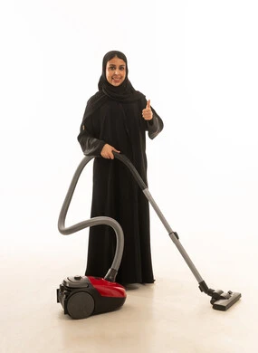 النظر للكاميرا مع رفع اصبع الابهام علامة للاعجاب ,امرأة عربية سعودية خليجية  ترتدي عباءة سوداء  تقوم بالتنظيف باستخدام المكنسة الكهربائية بسعادة. , خلفية بيضاء