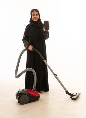 بورتريه لامرأة عربية سعودية خليجية بخلفية بيضاءترتدى عباءة خليجية سوداء  تنظر للكاميرا بابتسامة سرور تستخدم المكنسة الكهربائية مع عرض شاشة سوداء على الهاتف المحمول