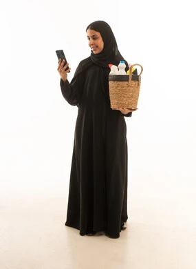 A portrait with a white background of a Saudi Gulf woman wearing a black Gulf abaya, carrying cleaning and household supplies, using her mobile phone to purchase household necessities.