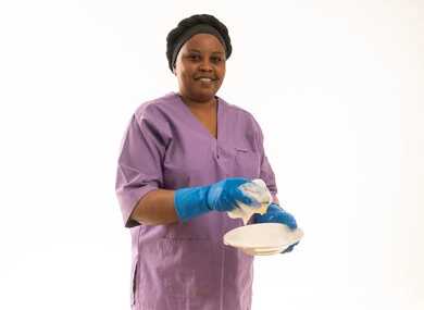 بورتريه لعاملة منزلية ترتدى يونيفورم باللون البنفسجى تقوم بغسل الصحون بالصابون ,شغالة اقريقية ترتدي قفاز أزرق تهتم بنظافة المنزل ,ايماءات السعادة بالعمل ,خلفية بيضاء