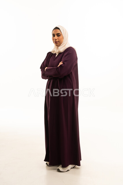 Looking at the camera with gestures of joy and self-confidence, the concept of elegance and attention to appearance, a portrait of a Gulf Arab Saudi woman wearing a hijab and traditional abaya standing with crossed arms, full-body image, white background.