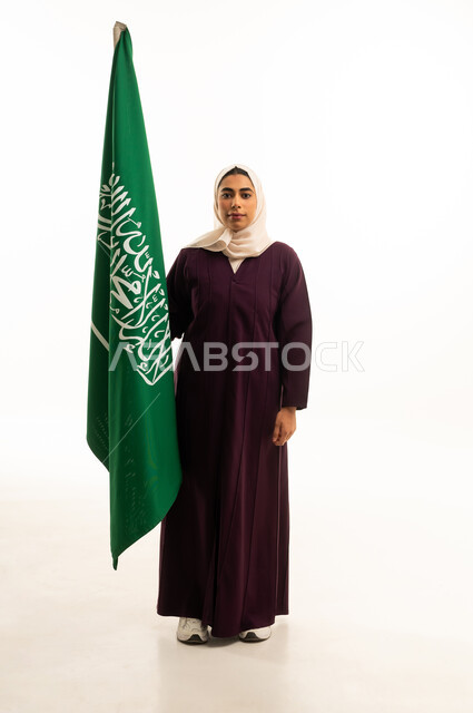 بورتريه لامرأة عربية خليجية سعودية محجبة ترتدي عباءة  تحمل العلم, الاحتفال بذكرى اليوم الوطني السعودي 8 ربيع الأول, الفخر و الاعتزاز بالانتماء للمملكة،  حب الوطن والانتماء إليه, الجسم بالطول الكامل, خلفية بيضاء