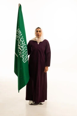 بورتريه لامرأة عربية خليجية سعودية محجبة ترتدي عباءة  تحمل العلم, الاحتفال بذكرى اليوم الوطني السعودي 8 ربيع الأول, الفخر و الاعتزاز بالانتماء للمملكة،  حب الوطن والانتماء إليه, الجسم بالطول الكامل, خلفية بيضاء
