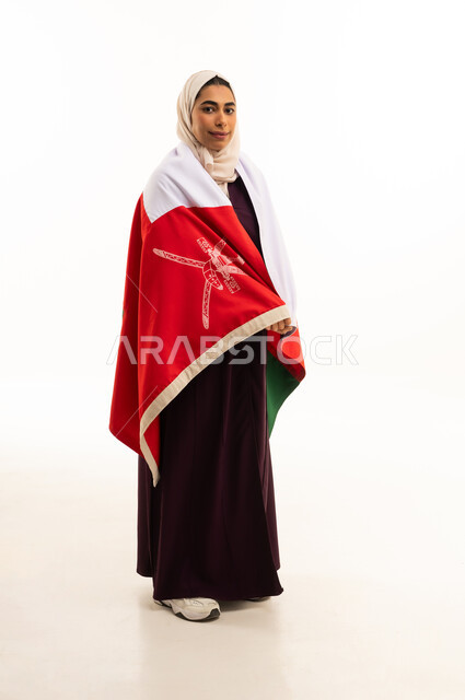 A portrait of an Arab Gulf Omani woman wearing a colorful abaya and a hijab, draping the national flag of the country over her shoulder, celebrating the anniversary of Omani National Day on November 18, love for the homeland and belonging to it, national symbols and cultures, commemorating the founding of the state in 1775 AD, full-body, white background.