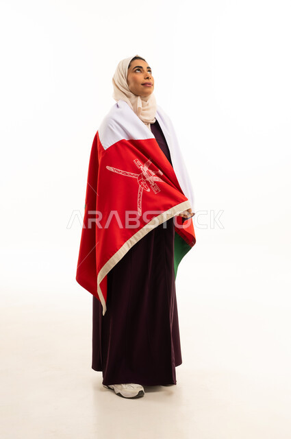 النظر الى شىء ما, بورتريه لإمرأة عربية خليجية عمانية محجبة ترتدي العباءة الملونة تضع العلم الوطني للبلاد على كتفها, الاحتفال بذكرى اليوم الوطني العماني 18 نوفمبر, احياء ذكرى تأسيس الدولة 1775 م , الجسم بالطول الكامل، خلفية بيضاء