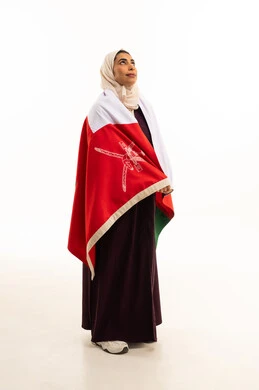 النظر الى شىء ما, بورتريه لإمرأة عربية خليجية عمانية محجبة ترتدي العباءة الملونة تضع العلم الوطني للبلاد على كتفها, الاحتفال بذكرى اليوم الوطني العماني 18 نوفمبر, احياء ذكرى تأسيس الدولة 1775 م , الجسم بالطول الكامل، خلفية بيضاء