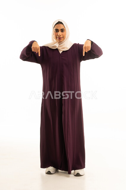 Confirmation of something happening at this moment, movements indicating this place (here), specifying a certain location, a portrait of a Saudi Arabian Gulf woman wearing a hijab and an abaya, pointing with her index finger downwards, looking at the camera with gestures of joy, full-body shot, white background.