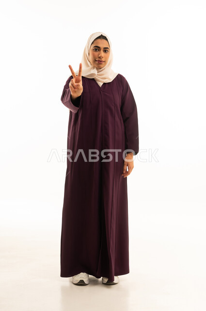 النجاح وتحقيق الأهداف, مفهوم السعادة بالوصول إلى الغايات, بورتريه لإمرأه عربيه خليجيه سعودية محجبه ترتدي العباءه ترفع يدها بإشارة النصر والقوة, إيماءات الفرح بالفوز والنصر, النظر إلى الكاميرا بتعابير السرور, الجسم بالطول الكامل, خلفية بيضاء