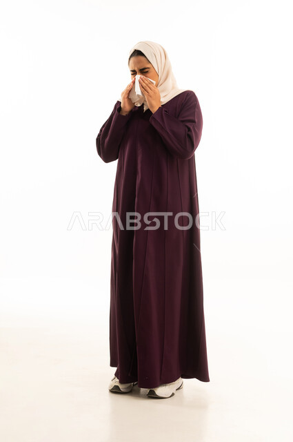 A side portrait of a Saudi Arabian Gulf woman wearing a hijab and an abaya, standing with expressions indicating sneezing and fatigue, suffering from a cold and flu, full body, white background.