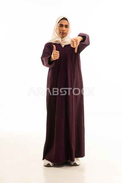 الموافقة وعدم الموافقة، بورتريه لامرأة عربيه خليجيه سعوديه مبتسمه محجبه ترتدي العباءة ترفع اصابع الابهام للاعلى وللاسفل بتعابيرالتردد في شيء ما, عدم الإعجاب و الرفض و القبول و الرضا, النظر الى الكاميرا بتعابير الحيرة، الجسم بالطول الكامل, خلفية بيضاء