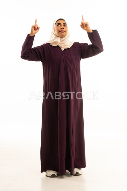 بورتريه لامرأة عربيه خليجيه سعوديه مبتسمه محجبه ترتدي العباءة وترفع اصابعها وتشير الى شيء ما, النظر الى الاعلى بإيماءات السرور، إشارات وحركات يدين تدل على هذا المكان (هنا)، تحديد موقع معين بإيماءات البشاشة، الجسم بالطول الكامل, خلفية بيضاء