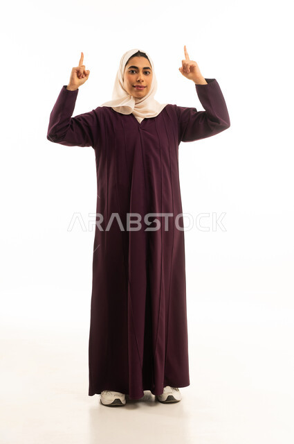 النظر الى الاعلى بإيماءات السرور، إشارات وحركات يدين تدل على هذا المكان (هنا)، تحديد موقع معين بإيماءات البشاشة، بورتريه لامرأة عربيه خليجيه سعوديه مبتسمه محجبه ترتدي العباءة وترفع اصابعها وتشير الى شيء ما,  الجسم بالطول الكامل, خلفية بيضاء