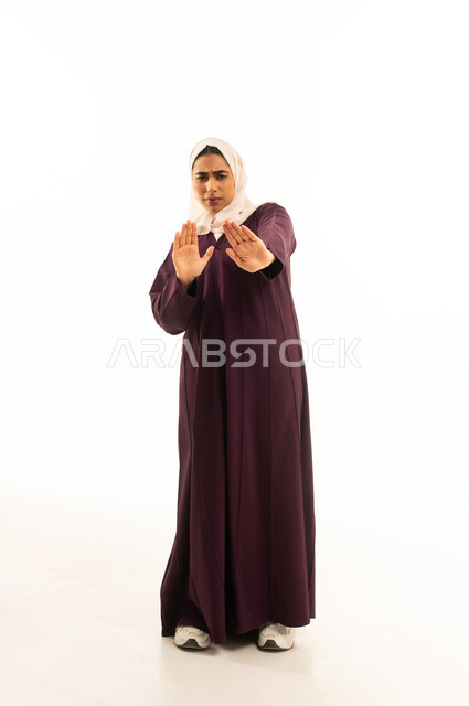 Protecting oneself from external harm, a portrait of a Saudi Arabian Gulf woman wearing a hijab and an abaya, raising her hands upwards with gestures and facial expressions indicating pause and awe, full-body, white background.