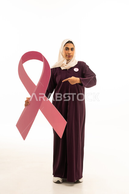 الاشارة الى شعار الحملة العالمية لسرطان الثدى, بورتريه لإمرأة عربية خليجية سعودية محجبة ترتدي العباءة تحمل شعاراليوم العالمي للتوعيه بالكشف المبكر ,الجسم بالطول الكامل, خلفية بيضاء