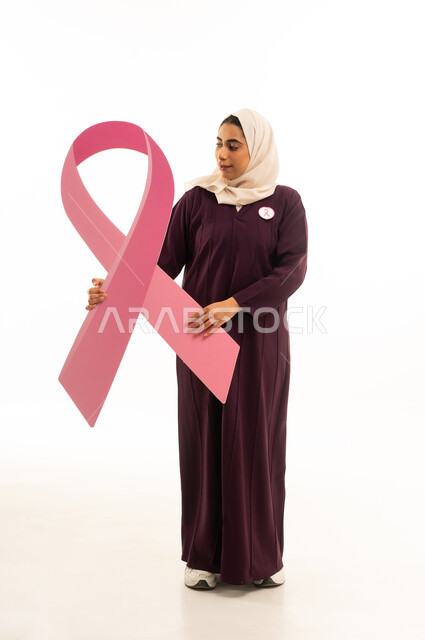 تحدي الأمراض الخبيثة, بورتريه لإمرأة عربية خليجية سعودية محجبة ترتدي العباءة تمسك شعار التوعيه بالكشف المبكر لسرطان الثدي, الجسم بالطول الكامل, خلفية بيضاء