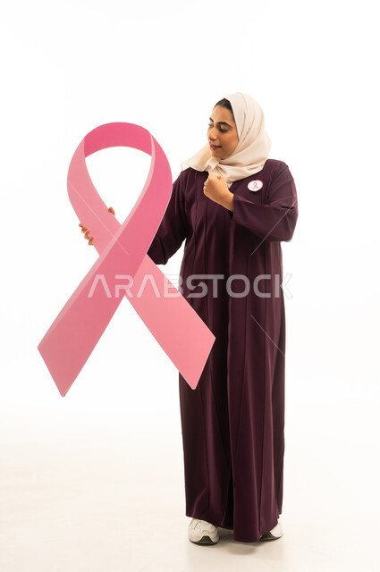 القوة والانتصار والتشجيع, بورتريه لإمرأة عربية خليجية سعودية محجبة ترتدي العباءة تمسك شعار التوعيه بالكشف المبكر لسرطان الثدي, الجسم بالطول الكامل, خلفية بيضاء
