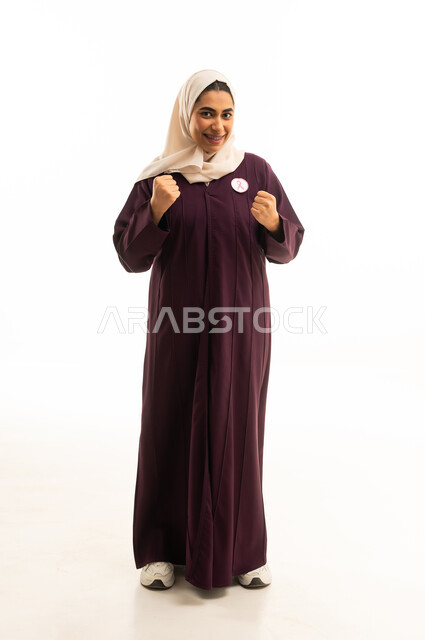Strength, victory, and encouragement, success and winning and achieving goals, a portrait of a smiling Arab Gulf Saudi woman wearing an abaya, raising her hands in gestures of enthusiasm, full-body, white background.
