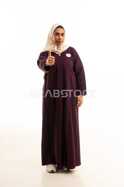 تعابير الموافقة و القبول ,النظر الى الكاميرا بإيماءات السرور, بورتريه لإمرأة عربية خليجية سعودية محجبة ترتدي العباءة و  شعار الحملة العالمية لسرطان ترفع اصبع الابهام للاعلى بعلامة الامتياز و الجودة , الجسم بالطول الكامل, خلفية بيضاء
