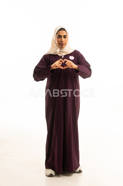 A hand gesture forming a heart of love sending affection, attention to appearance and elegance, expressions of joy and a smile, a portrait of a Saudi Arabian Gulf woman wearing a brown abaya, the concept of expression through gesture, full-body, white background.