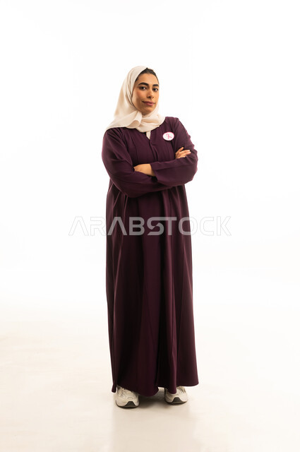 الوقوف بأيدى متقاطعة، الاناقة والاهتمام بالظهر الخارجي, بورتريه لامرأة عربية خليجية سعودية مبتسمة محجبة ترتدي العباءة البنية  و شعار التوعيه بالكشف المبكر لسرطان الثدي، الحملة التوعويه بالمرض الخبيث، الجسم بالطول الكامل, خلفية بيضاء