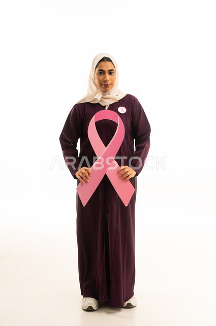 اليوم العالمي للتوعية بالمرض الخبيث بورتريه لامرأة عربية خليجية سعودية مبتسمة محجبة ترتدي العباءة البنية تمسك شعار التوعيه بالكشف المبكر لسرطان الثدي، الحملة التوعويه بالمرض الخبيث، الجسم بالطول الكامل, خلفية بيضاء