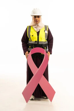 Looking at the camera with expressions of self-confidence, World Cancer Awareness Day, a portrait of a smiling Saudi Gulf Arab engineer wearing a safety vest and helmet holding in her hands the awareness slogan for early detection of breast cancer, full body, white background.