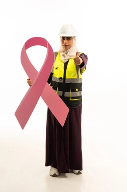 النظر إلى الكاميرا بإشارة الموافقه, بورتريه لمهندسة عربية خليجية سعودية مبتسمة ترتدي سترة الحمايه مع الخوذه تمسك بيديها شعار التوعيه بالكشف المبكر لسرطان الثدي ترفع يدها بإيماءات علامة الجودة و السرور, الجسم بالطول الكامل, خلفية بيضاء