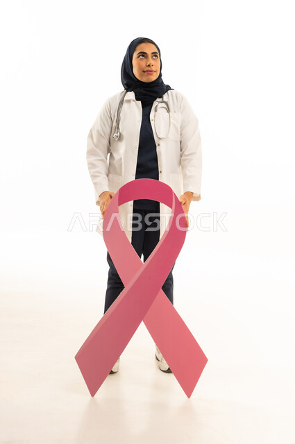 النظر الى شىء ما فى الاعلى, بورتريه لطبيبة عربية خليجية سعودية محجبة ترتدي البالطو الابيض و السماعة الطبية تحمل الشعار الوردى للتوعيه بالكشف المبكر لسرطان الثدى, الجسم بالطول الكامل, خلفية بيضاء