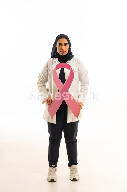 النظر إلى الكاميرا بتعابير الثقة بالنفس,  بوتريه لامرأة عربية خليجية سعودية محجبة ترتدي الزى الكاجوال تمسك بيديها شعارالحملة العالمية الوردى لمكافحة سرطان الثدى بإيماءات الانتصار والنجاح, خلفية بيضاء