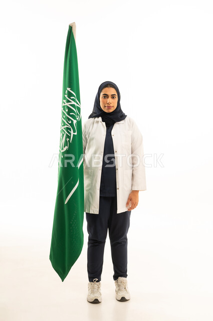 العيد الوطني 23 سبتمبر, مفهوم حب الوطن والانتماء له,  يوم العلم السعودي 11 مارس، بورتريه لامرأة عربية خليجية سعودية محجبة ترتدي الزى الكاجوال تنظرالى علم المملكة, المشاركة بالأعياد الوطنية، الجسم بالطول الكامل, خلفية بيضاء