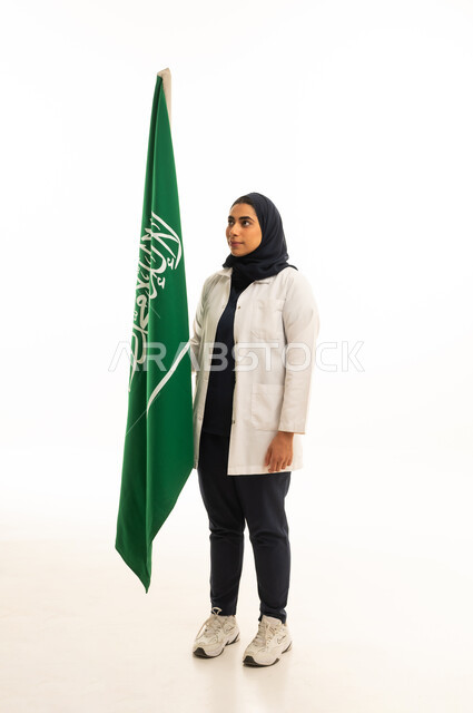 بورتريه من الجانب لامرأة عربية خليجية سعودية محجبة ترتدي الزى الكاجوال تحمل علم المملكة, المشاركة بالأعياد الوطنية، يوم العلم السعودي 11 مارس، العيد الوطني 23 سبتمبر، الجسم بالطول الكامل, خلفية بيضاء