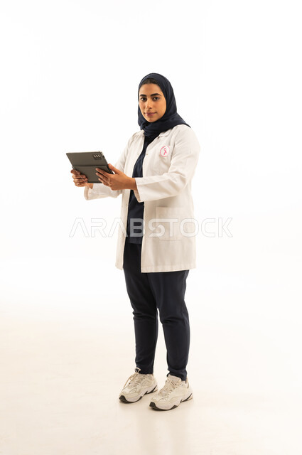 النظر الى الكاميرا بتعابير الثقة, بورتريه لامرأة عربية خليجية سعودية محجبة ترتدي الزى الكاجوال و شعار التوعيه بالكشف المبكر لسرطان الثدي تحمل التابلت بيدها، اليوم العالمي للتوعية بالامراض الخبيثة, الجسم بالطول الكامل, خلفية بيضاء