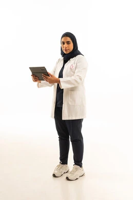 النظر الى الكاميرا بتعابير الثقة, بورتريه لامرأة عربية خليجية سعودية محجبة ترتدي الزى الكاجوال و شعار التوعيه بالكشف المبكر لسرطان الثدي تحمل التابلت بيدها، اليوم العالمي للتوعية بالامراض الخبيثة, الجسم بالطول الكامل, خلفية بيضاء