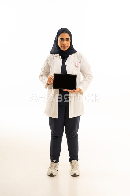 بورتريه لامرأة عربية خليجية سعودية محجبة ترتدي الزى الكاجوال و شعار التوعيه بالكشف المبكر لسرطان الثدي تعرض شاشة سوداء عبر التابلت، اليوم العالمي للتوعية بالامراض الخبيثة, الجسم بالطول الكامل, خلفية بيضاء