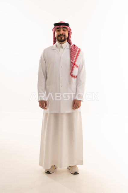نظرة أمامية للكاميرا , خلفية بيضاء , الجسم بالطول الكامل , طبيب عربي خليجي سعودي يرتدي الزي التقليدي في المملكة مع المعطف الطبي الابيض , ايماءات السعادة