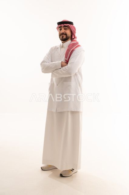 A Saudi Arabian Gulf Arab doctor wearing a white medical coat over the traditional attire of the kingdom stands proudly with his arms crossed, against a white background, full body.